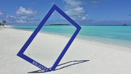 Fototapeta premium There is a blue photo frame on the white sand of the beach. The aquamarine ocean is calm. A leaning palm tree, a wooden footpath, a pier are visible in the distance. Maldives.