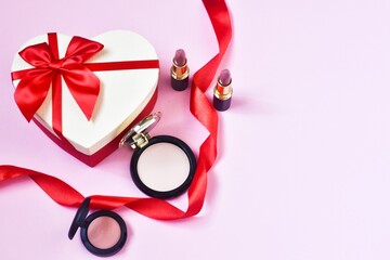 Gift box in the form of a heart with decorative cosmetics, with red ribbon on a pink background. Concept beauty, holiday. Copy space. Flat lay.