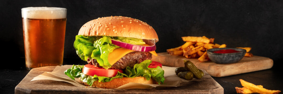 Burger and beer panorama. Hamburger with beef, cheese, onion, tomato, and green salad, with pickles and French fries, a side view on a dark background