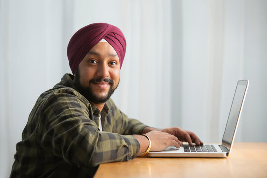 A SIKH MAN LOOKING AT CAMERA AND SMILING WHILE AT WORK WITH LAPTOP	