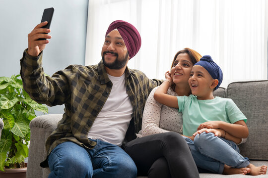 A SIKH SON HUGS MOTHER WHILE FATHER CLICKS A SELFIE OF EVERYONE POSING TOGETHER ON A SOFA	