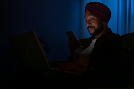 A SIIKH MAN CHECKING MOBILE PHONE WHILE WORKING ON LAPTOP	