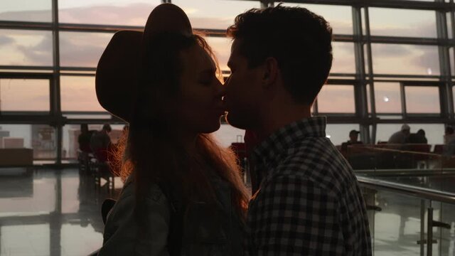 Silhouette of man and a woman kissing in airport terminal after arrival. Concept of travel. Happy couple meet after long separation. Sunset panoramic window at background. 6k downscale 10 bit.