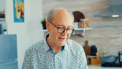 Cheerful senior man during video conference in kitchen while enjoying breakfast and a cup of coffee. Elderly person using internet online chat technology video webcam making a video call connection