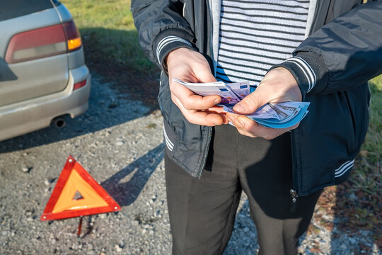 Money In The Hands Of A Person Who Counts It Against The Background Of A Car Accident, An Emergency Sign And The Roadside