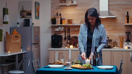 Young woman lighting the candle waiting her husband for a romantic dinner. Caucasian happy wife preparing festive meal with healty food for anniversary celebration, sitting near the table in kitchen.