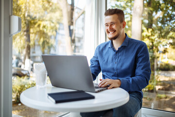 Fototapeta premium Cheerful businessman in a blue shirt working from a cafe on his laptop. Young man leads distance business with a cup of coffee. Remote job concept.
