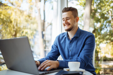 Cheerful businessman in a blue shirt working from a cafe on his laptop. Young man leads distance business with a cup of coffee. Remote job concept.