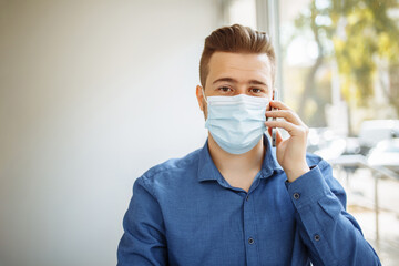 Young businessman wearing a medical sterile mask talks on the mobile phone leading business from a cafe, working remotely during coronavirus pandemic quarantine. Health safety concept.