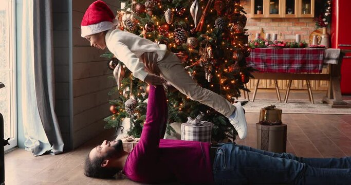 African ethnicity father lying on warm wooden floor in decorated with lights and Christmas tree living room holding lifting up preschool son, family having fun playing at home, happy holidays concept