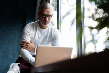 Fototapeta premium Concentrated on work. Casual Grey-haired Mature man in eyeglasses working on laptop while sitting in office or cafe near the window