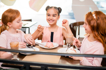 Three little girls sitting at table in a cafe cheerfully drink hot chocolate and eat cakes.
