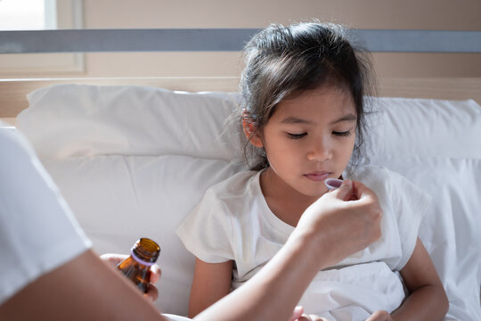 Asian Mother Giving Cough Syrup Medicine On A Spoon To Her Daughter In Bed, Sick Child And Health Care Concept.