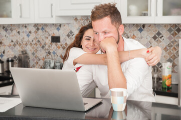 happy couple woman and confused man sitting at kitchen with laptop thinking and hugging