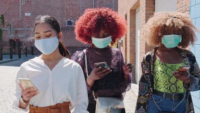 group of three young multiethnic female people walking down the street wearing a face mask as protection against covid-19 and holding mobile phones. New normal and social gathering concept