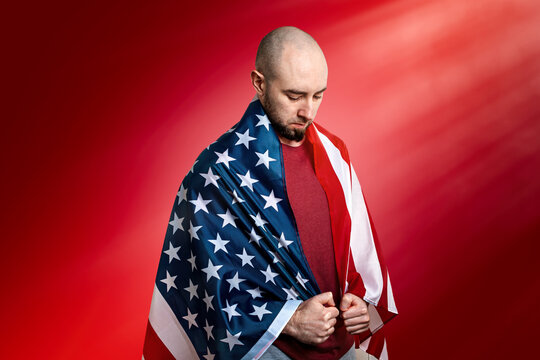 A Caucasian Man Covered With An American Flag Stands In A Humble And Proud Pose. Red Background. Copy Space. Concept Of Memorial Day, Independence Day, And Other National Holidays In The United States