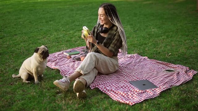 Smiling girl taking photo with cute pug puppy in green city park holding camera, sitting on plaid