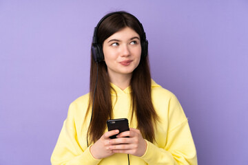 Young Ukrainian teenager girl over isolated purple background listening music with a mobile and thinking