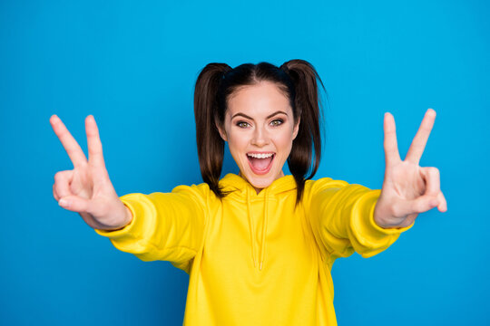 Photo of pretty funny lady two tails hairdo raise both arms showing v-sign symbol good cheerful mood say hi friends wear casual yellow hoodie pullover isolated bright blue color background