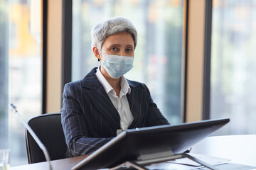 Portrait of mature businesswoman in protective mask sitting at the table with computer and looking at camera at office