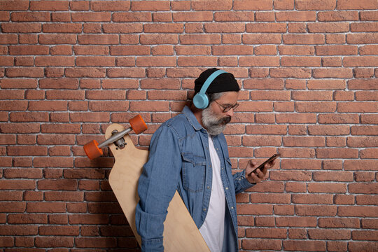 Profile view of modern man with beard, hat, headphones and longboard looking at phone on brick background