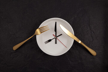 White deep plate, knife and fork. Clock on a dark textured stone background with copy space. The hands point to 8 o'clock. Concept - interval fasting or autophagy