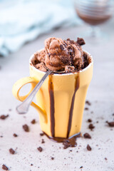 Chocolate brown ice cream in a regular yellow mug and spoon, on a blue background, with topping and crumbs. Unusual serve of dessert, copy space.