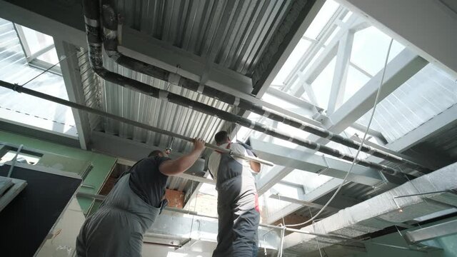 HVAC Installers On Scaffold Assembling Air Ventilation Shaft In Silver Material Hanging From Ceiling Inside New Building.