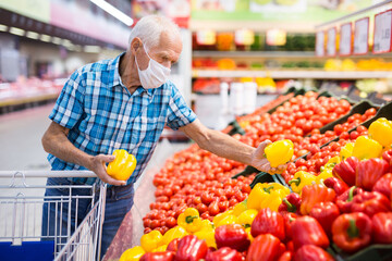 elderly caucasian man in mask with covid protection choosing tomatoes in vegetable section of supermarket