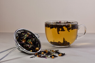 A glass cup with a hot herbal drink, a metal strainer with dry tea on a light, beige wooden background. Concept for tea drinking. Close up.