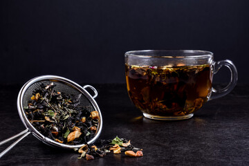 Glass cup with hot brewed tea, metal strainer with dry mix of herbs and fruits on a black wooden background. Concept for tea time.
