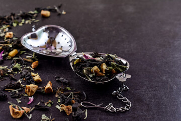 Steel, open filter, heart-shaped tea strainer, a mixture of herbs and fruits scattered on the table. Black, dark background. Close-up. Diagonal. Place for your text.