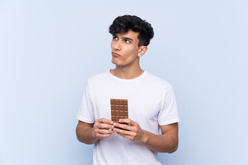 Young Argentinian man over isolated blue background taking a chocolate tablet and having doubts