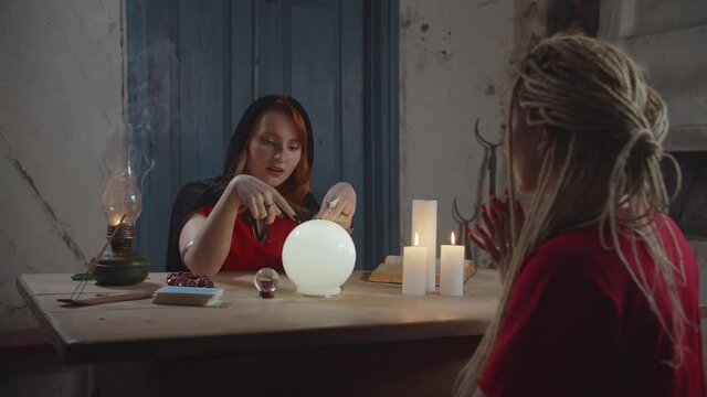 Mysterious pretty redhead soothsayer seeing visions of future events using crystal ball, fortune telling disappointing news to lovely woman while practicing divination in rural house at dusk.