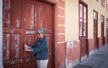 One senior caucasian woman with gray hair tries to open an old red door closed by lock. Traditional spanish home with red windows and doors made by wood. Rustic style.