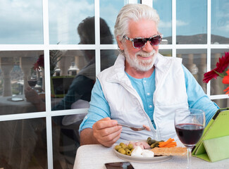 A smiling bearded senior man enjoying a light break eating and drinking while sitting at table on balcony using tablet. Concept of modern and tech retired people