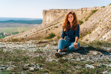 Naklejka na ściany i meble Beautiful young brunette woman in ripped jeans sitting on a cliff overlooking the rustic landscape, White rock in Crimea