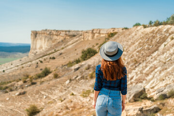 Fototapeta na wymiar Rear view of a young woman in a plaid shirt and hat standing on a mountain and admiring the landscape, White rock in the Crimea