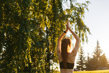 Rear view of flexible young woman raising hands up outside in city park. Back view of female practicing yoga outdoors in sunny summer morning.