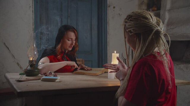 Beautiful red haired hand reader practicing chiromancy, fortune telling through study of female palm, interpreting future with ancient spellbook while doing divination in rustic house in the evening.