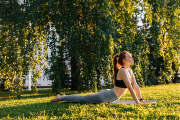 Side view of flexible young woman lying on yoga exercise mat and stretching neck and spine outside in city park background of sunray and green grass.