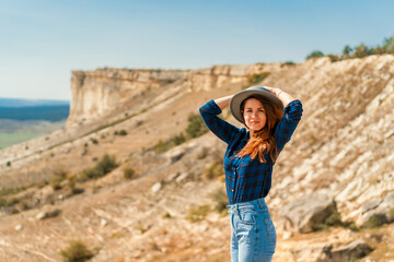 Naklejka premium A young woman in a plaid shirt and hat stands on a mountain with a view of the rustic landscape, White rock in the Crimea