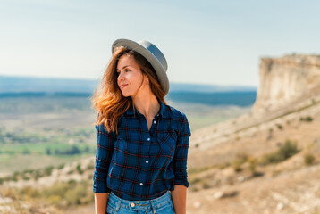 A young woman in a plaid shirt and hat stands on a mountain with a view of the rustic landscape, White rock in the Crimea