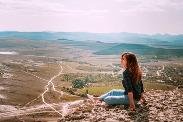 Beautiful young brunette woman in ripped jeans sitting on a cliff overlooking the rustic landscape, White rock in Crimea
