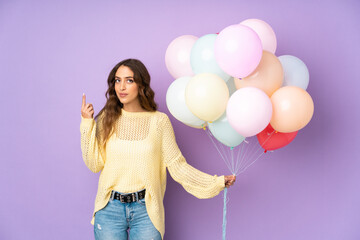 Young woman catching many balloons over isolated on purple background pointing with the index finger a great idea