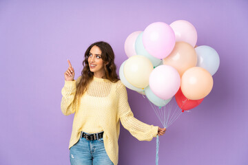 Young woman catching many balloons over isolated on purple background pointing with the index finger a great idea