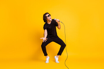 Fototapeta na wymiar Full length photo of young man dance hold mic sing open mouth wear black t-shirt pants white sneakers sunglass isolated yellow color background