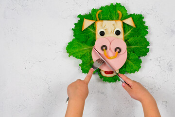 Funny sandwich with edible bull symbol of 2021 made of toast and sausage on green lettuce leaves. Children's hands are holding a fork and knife. Breakfast idea for children. New Year, Christmas food.