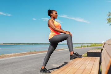 fitness, sport and healthy lifestyle concept - young african american woman stretching leg on bench at seaside