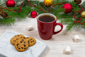 Red cup with tea, coffee with cookies and marshmallows on a white wooden table against the background of a New Year tree with Christmas decorations. Coziness and New Year concept.